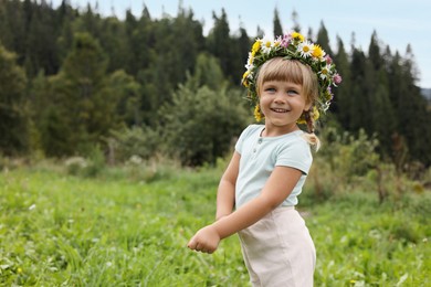 Portrait of smiling little girl in floral wreath at green meadow, space for text. Child enjoying beautiful nature