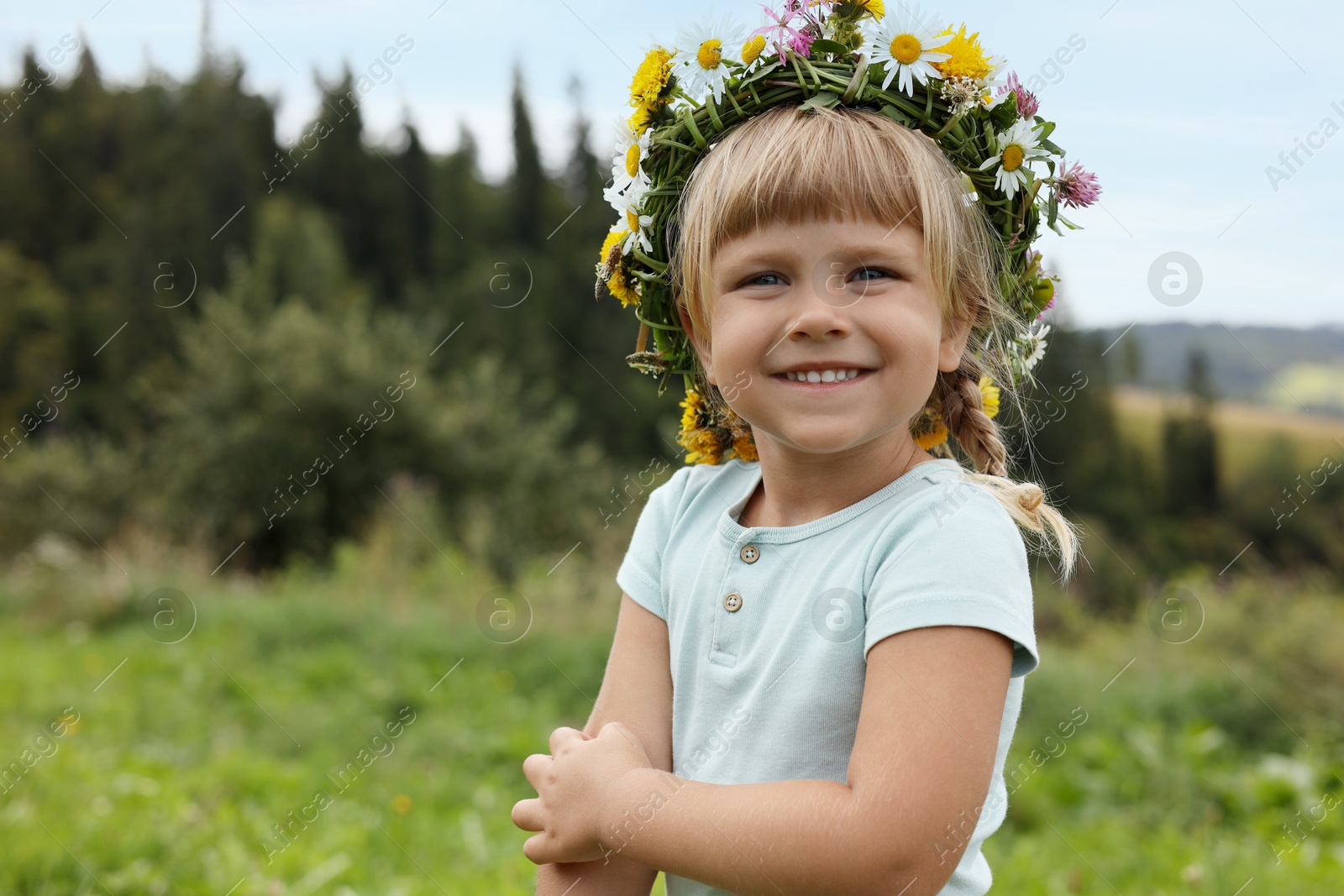 Photo of Portrait of smiling little girl in floral wreath outdoors, space for text. Child enjoying beautiful nature