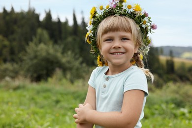 Photo of Portrait of smiling little girl in floral wreath outdoors, space for text. Child enjoying beautiful nature