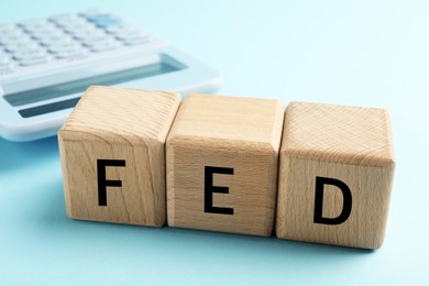 Photo of Wooden cubes with letters Fed (Federal Reserve System) and calculator on light blue background, closeup