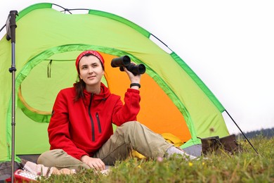 Young woman with binoculars sitting in tent on green grass outdoors, low angle view