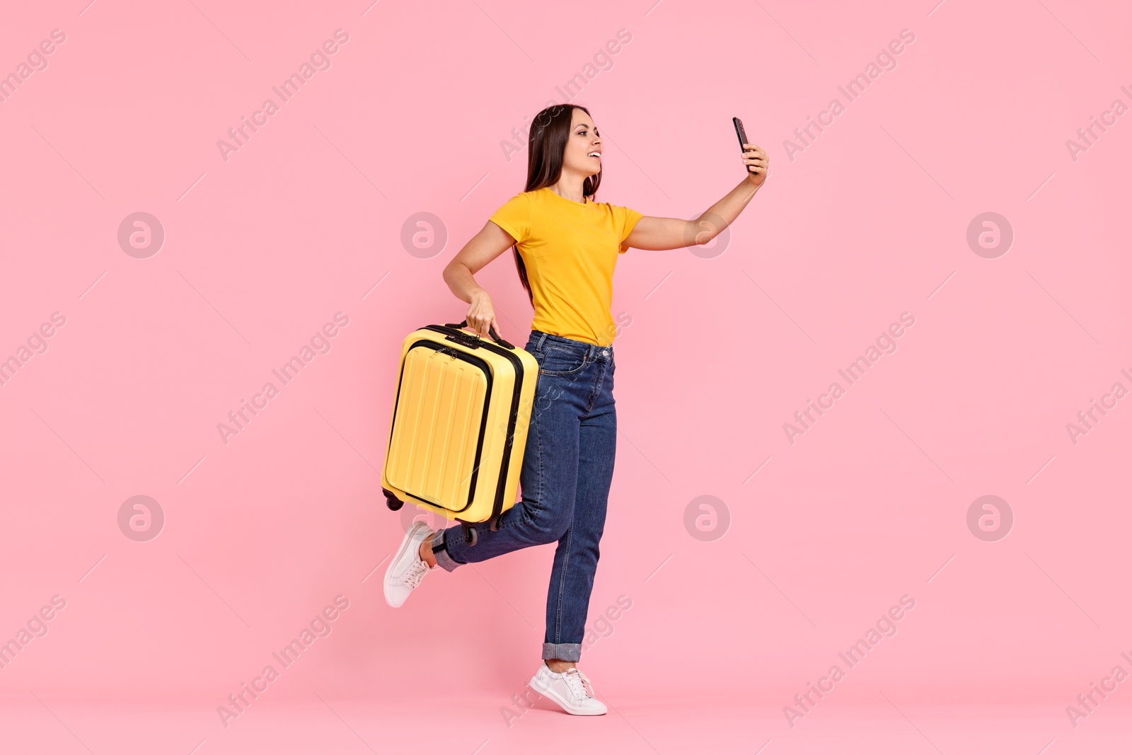 Photo of Happy young woman with suitcase taking selfie on pink background