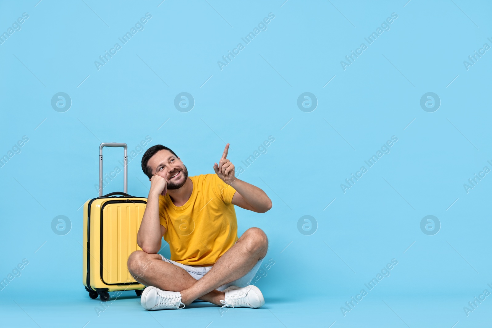 Photo of Happy man with suitcase pointing at something on light blue background, space for text