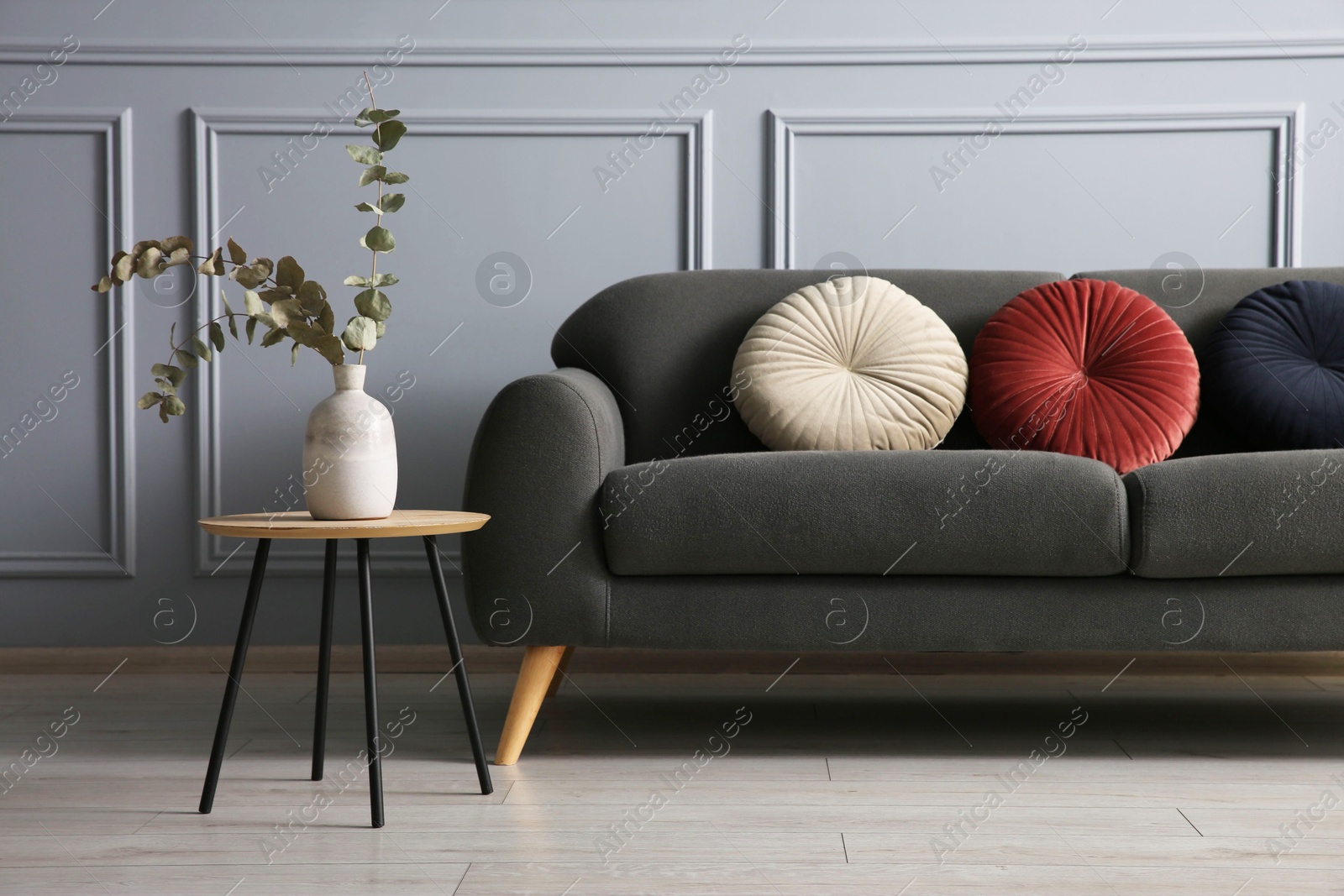 Photo of Stylish sofa with cushions and eucalyptus branches in vase on side table near grey wall