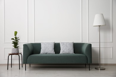 Photo of Stylish sofa with cushions, floor lamp and plant on side table near white wall