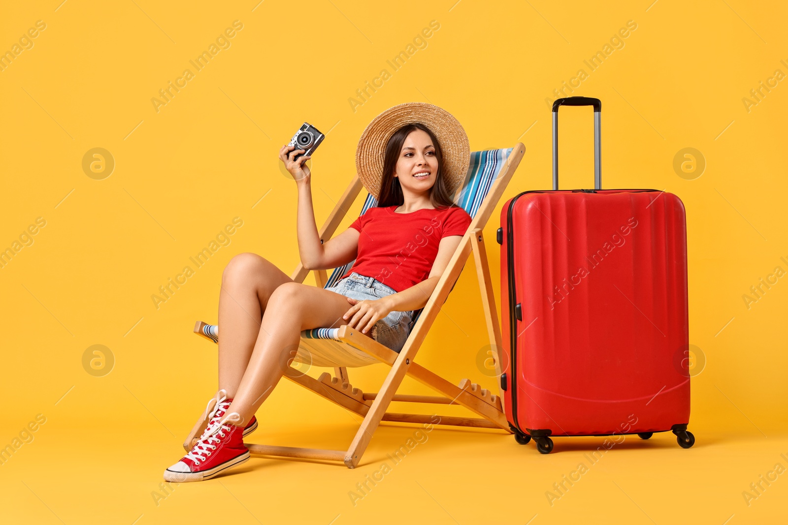 Photo of Happy young woman with vintage camera sitting on folding chair and suitcase against orange background