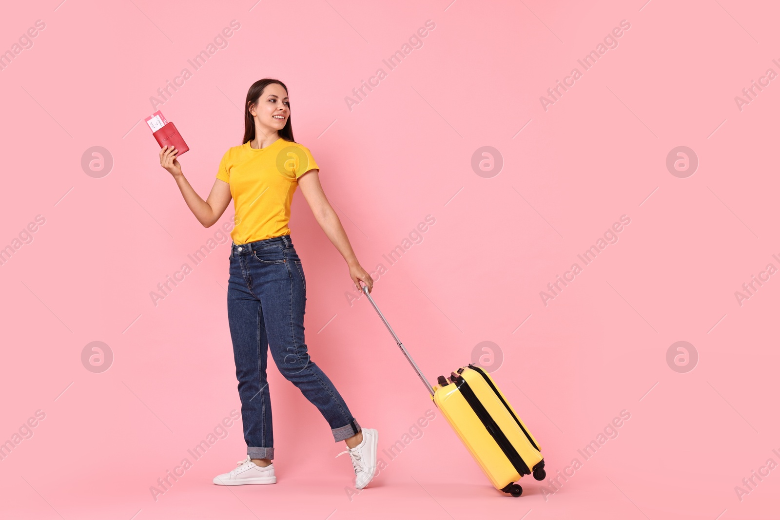 Photo of Happy young woman with suitcase, passport and ticket walking on pink background, space for text