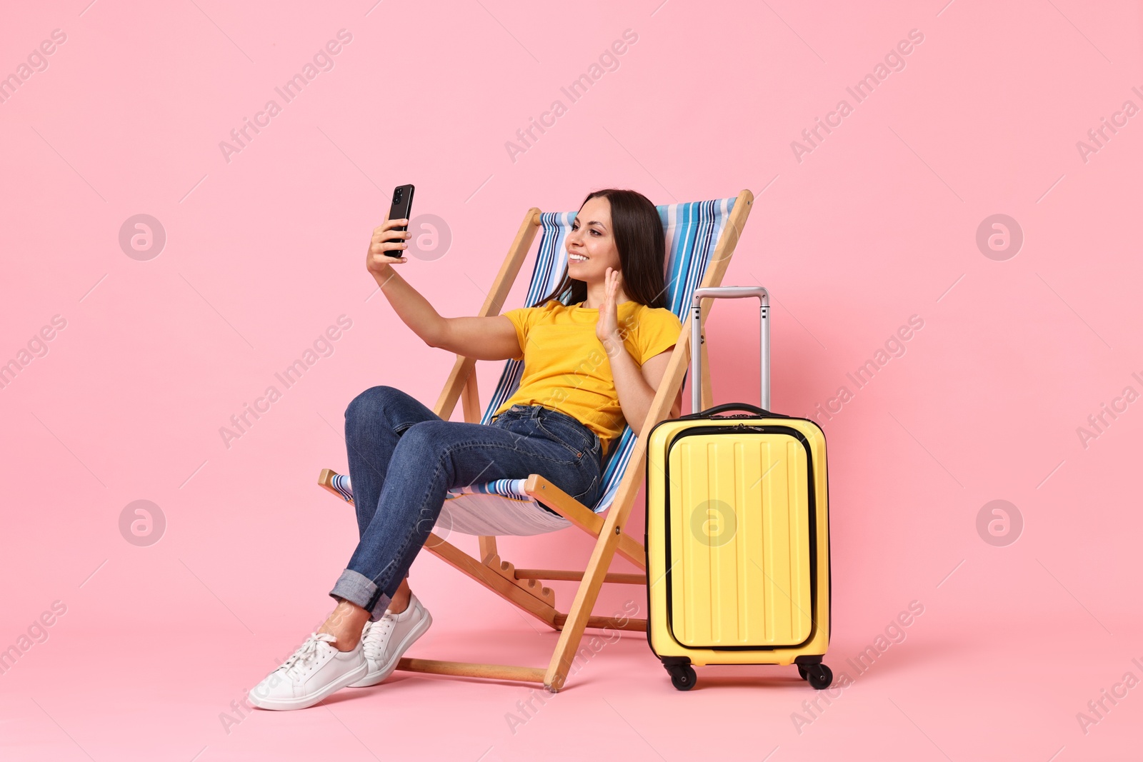 Photo of Happy young woman with smartphone greeting someone while sitting on folding chair near suitcase against pink background