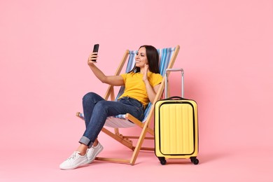 Photo of Happy young woman with smartphone greeting someone while sitting on folding chair near suitcase against pink background