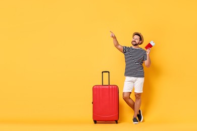 Photo of Happy man with suitcase, passport and ticket pointing at something on orange background