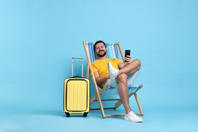 Happy man with smartphone sitting on folding chair and suitcase against light blue background