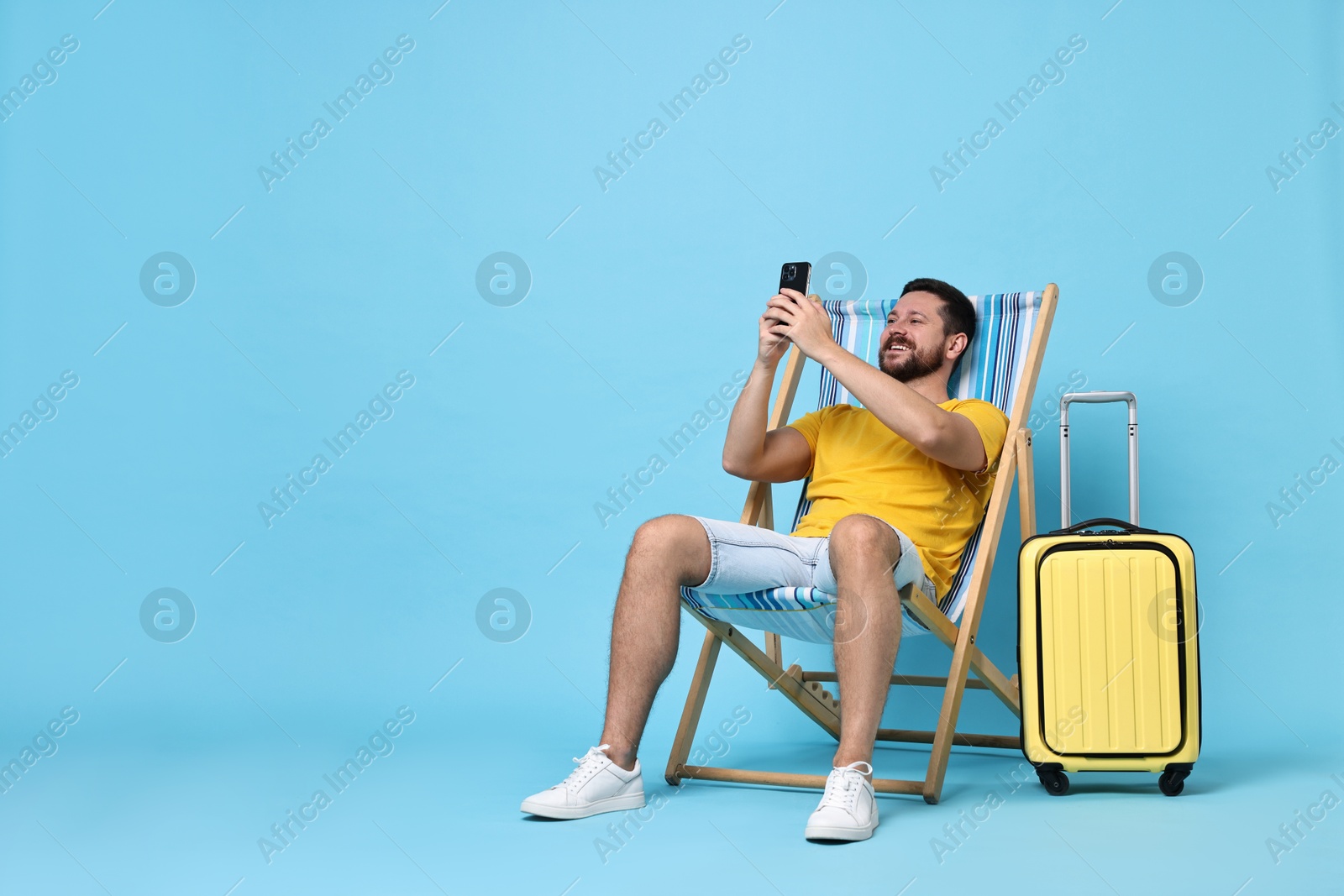 Photo of Happy man using smartphone while sitting on folding chair and suitcase against light blue background