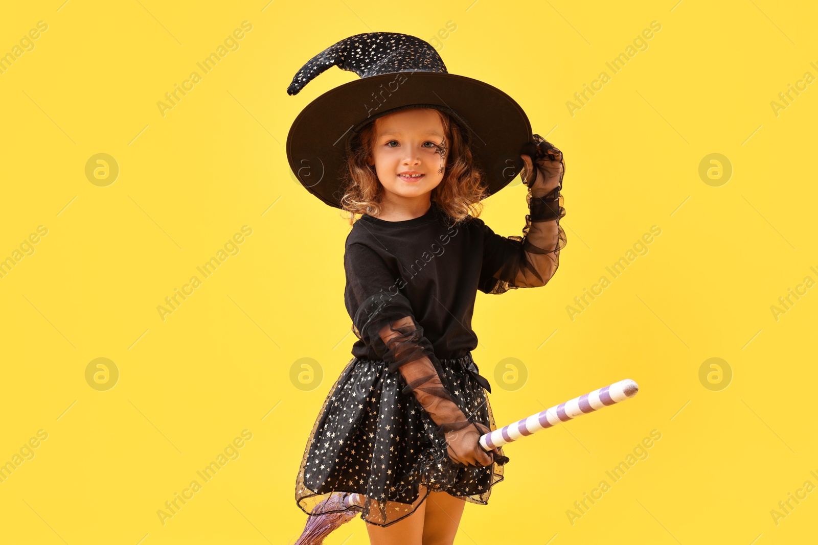 Photo of Cute girl with broom dressed like witch on yellow background. Halloween celebration