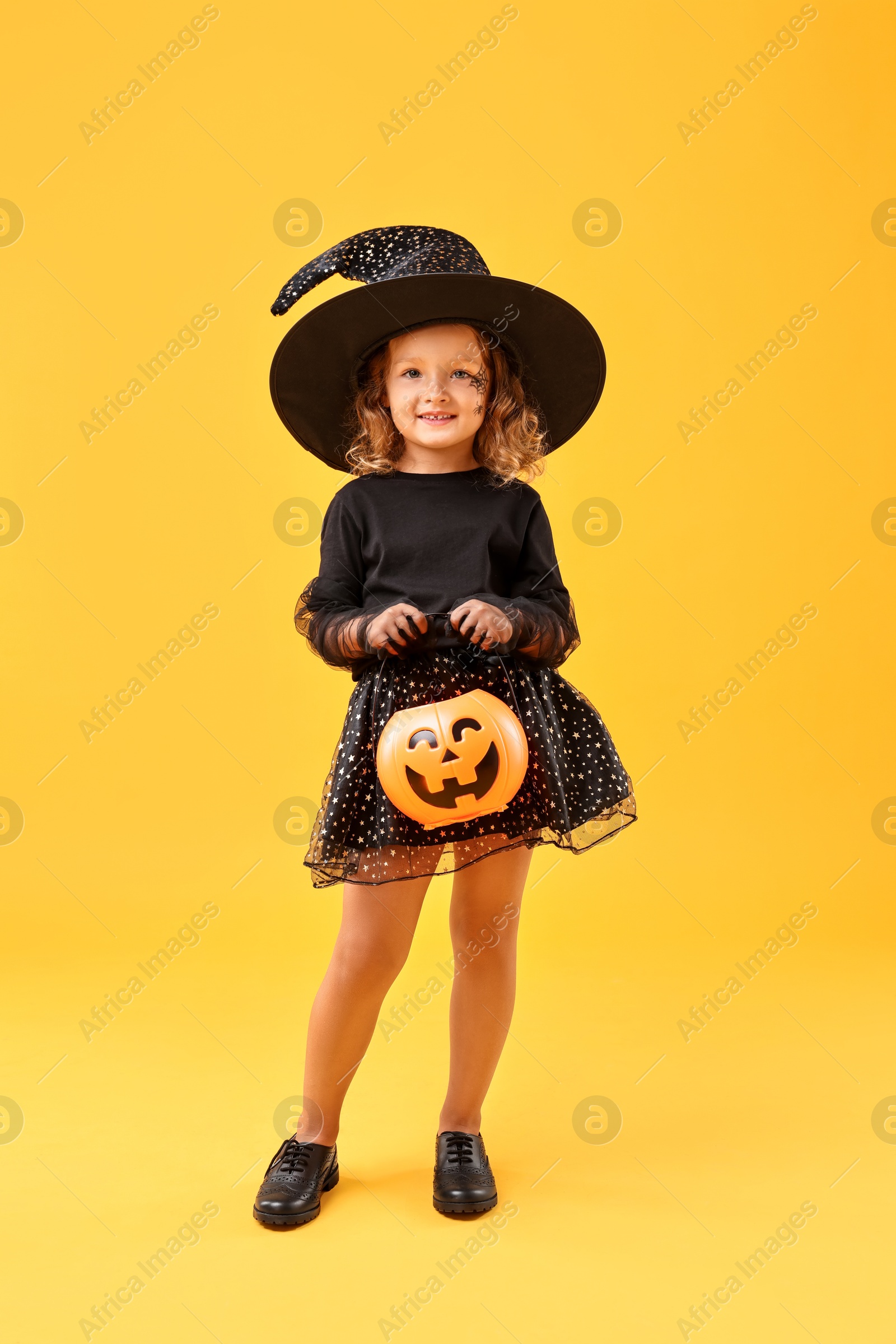 Photo of Cute girl with pumpkin bucket dressed like witch on yellow background. Halloween celebration