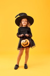 Photo of Cute girl with pumpkin bucket dressed like witch on yellow background. Halloween celebration