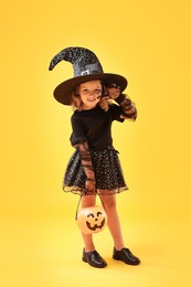 Cute girl with pumpkin bucket dressed like witch on yellow background. Halloween celebration