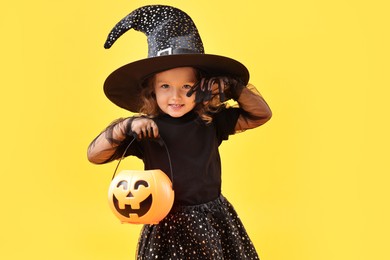 Photo of Cute girl with pumpkin bucket dressed like witch on yellow background. Halloween celebration