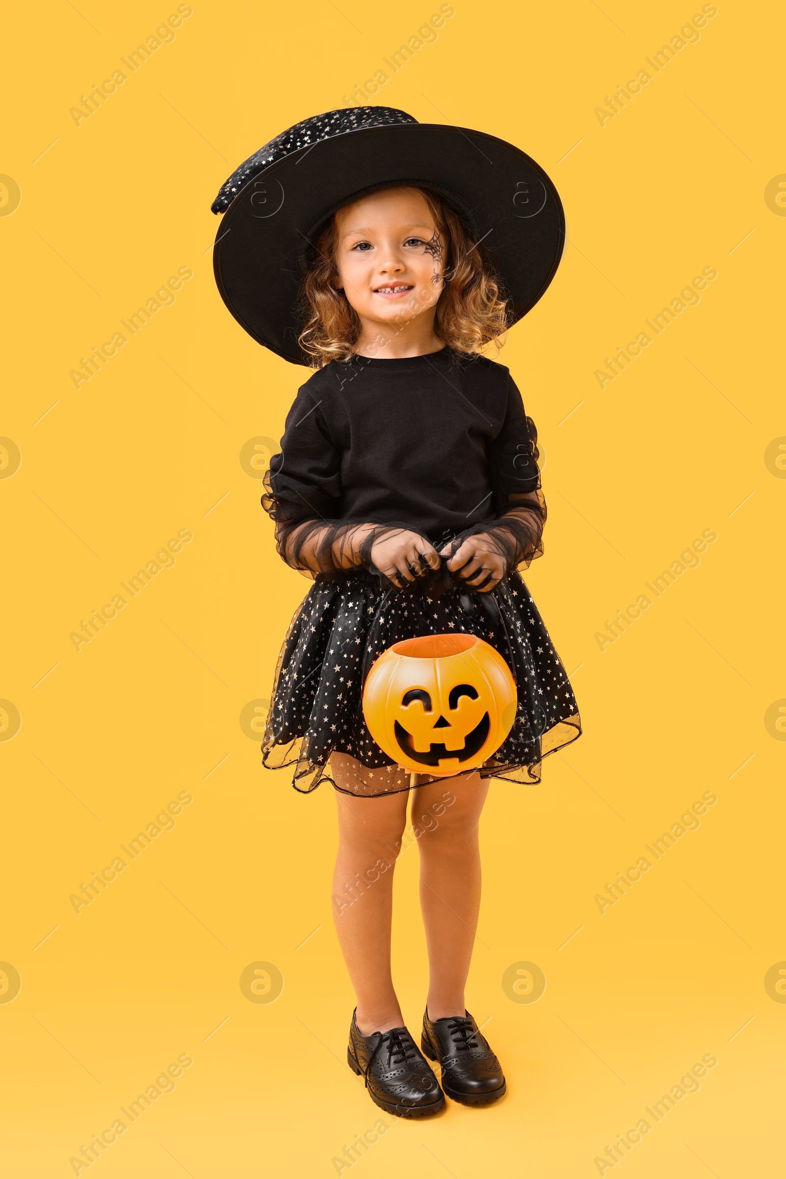 Photo of Cute girl with pumpkin bucket dressed like witch on yellow background. Halloween celebration