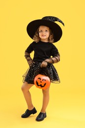 Photo of Cute girl with pumpkin bucket dressed like witch on yellow background. Halloween celebration