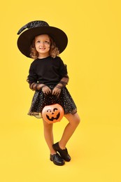 Photo of Cute girl with pumpkin bucket dressed like witch on yellow background. Halloween celebration