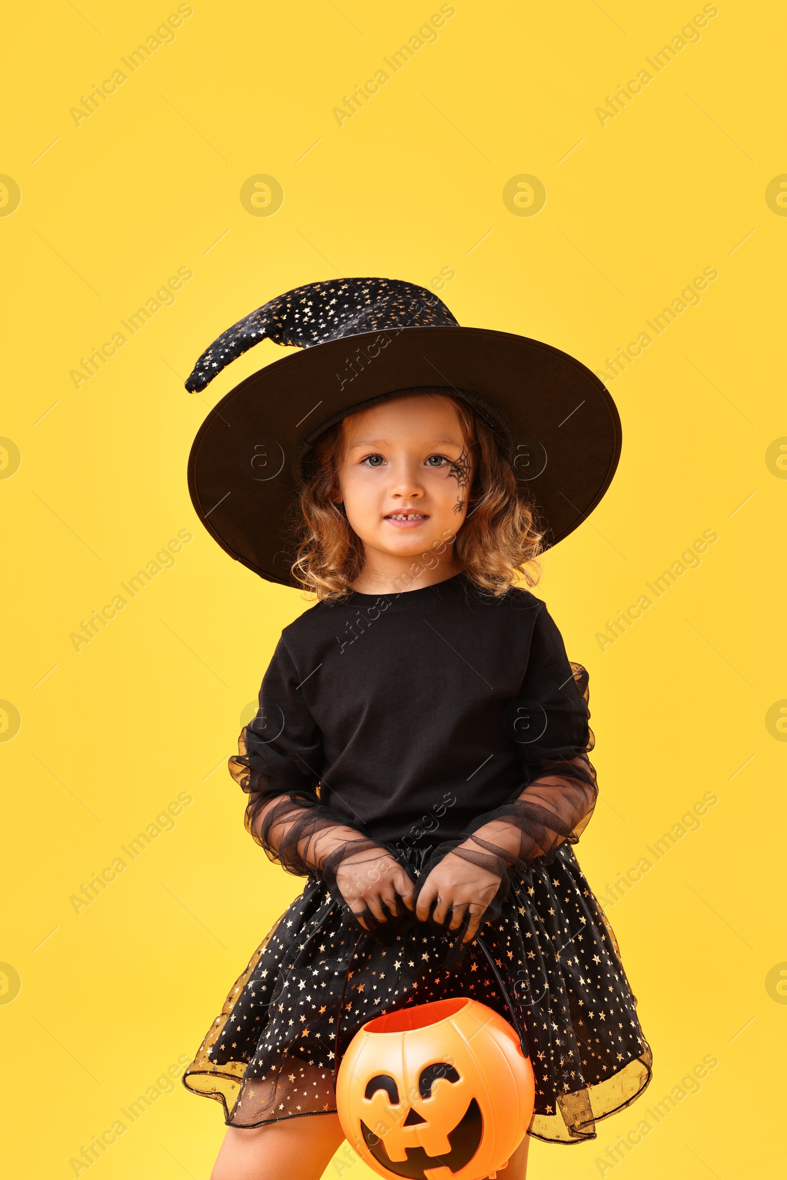 Photo of Cute girl with pumpkin bucket dressed like witch on yellow background. Halloween celebration