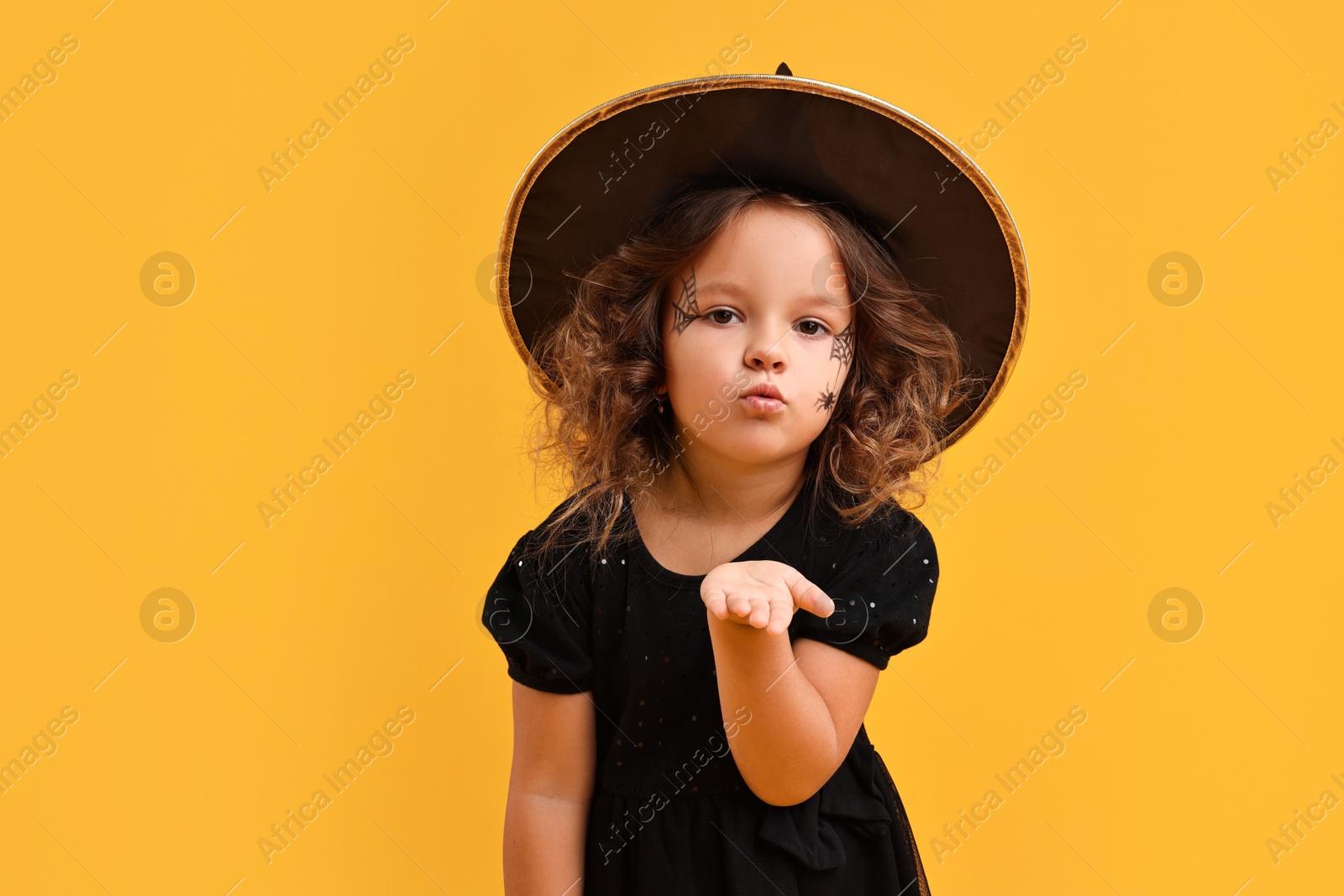 Photo of Cute girl dressed like witch blowing kiss on yellow background. Halloween celebration