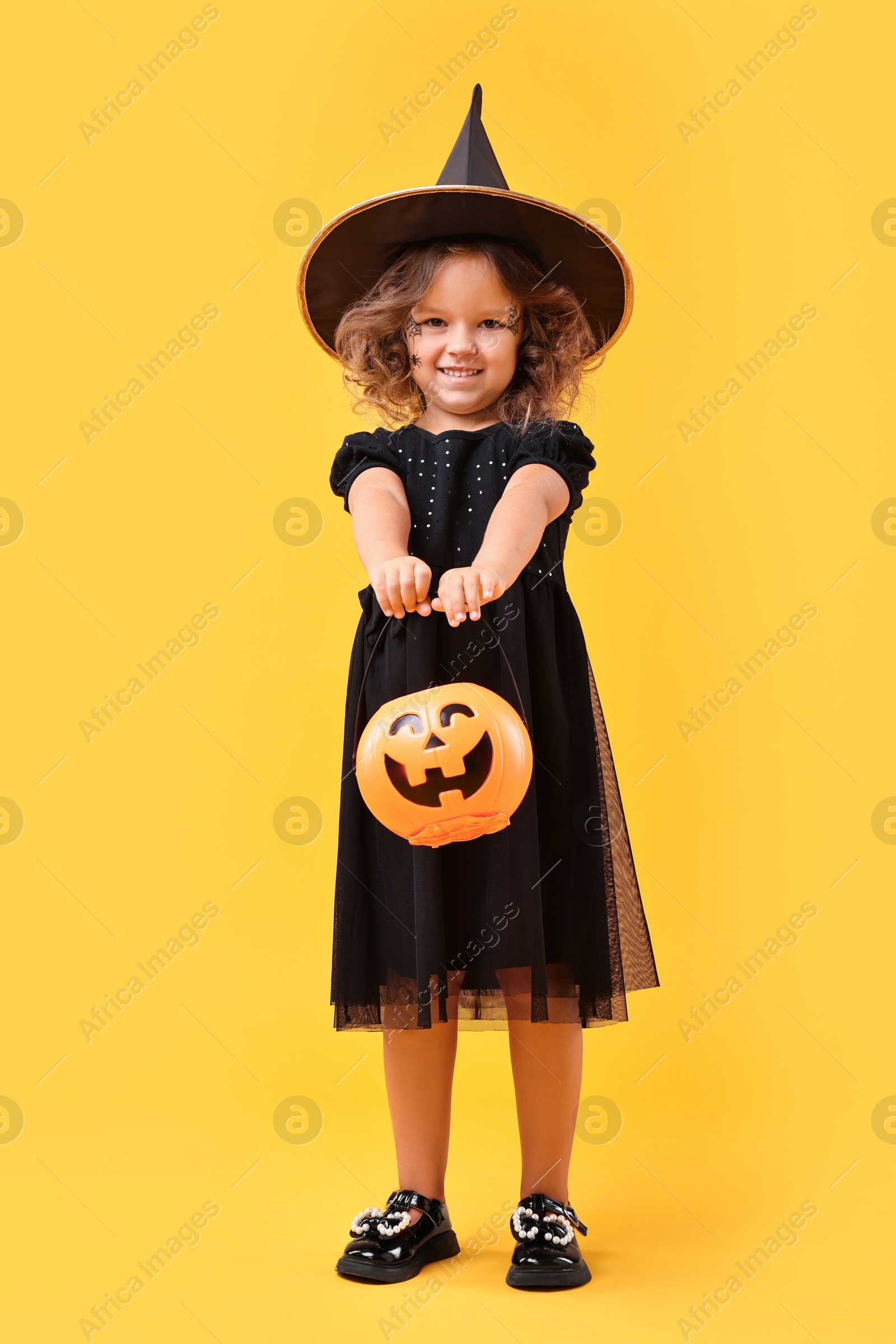 Photo of Cute girl with pumpkin bucket dressed like witch for Halloween celebration on yellow background