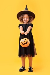 Photo of Cute girl with pumpkin bucket dressed like witch for Halloween celebration on yellow background