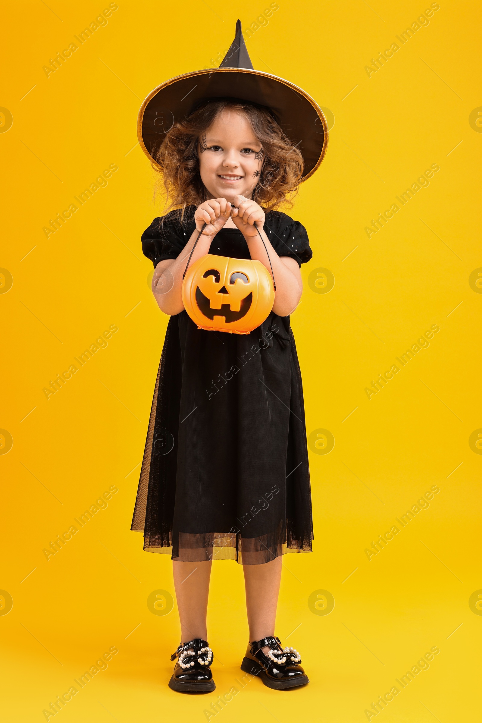 Photo of Cute girl with pumpkin bucket dressed like witch for Halloween celebration on yellow background