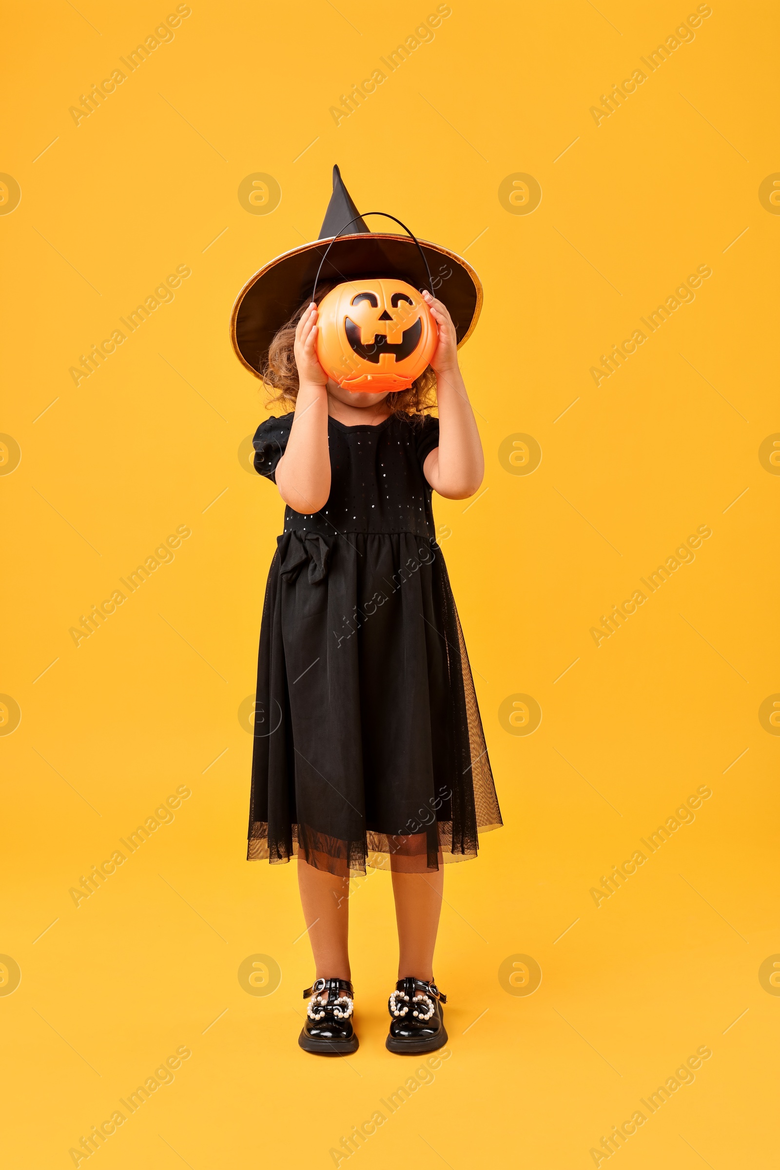 Photo of Girl with pumpkin bucket dressed like witch on yellow background. Halloween celebration