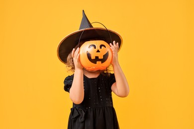 Photo of Girl with pumpkin bucket dressed like witch on yellow background. Halloween celebration