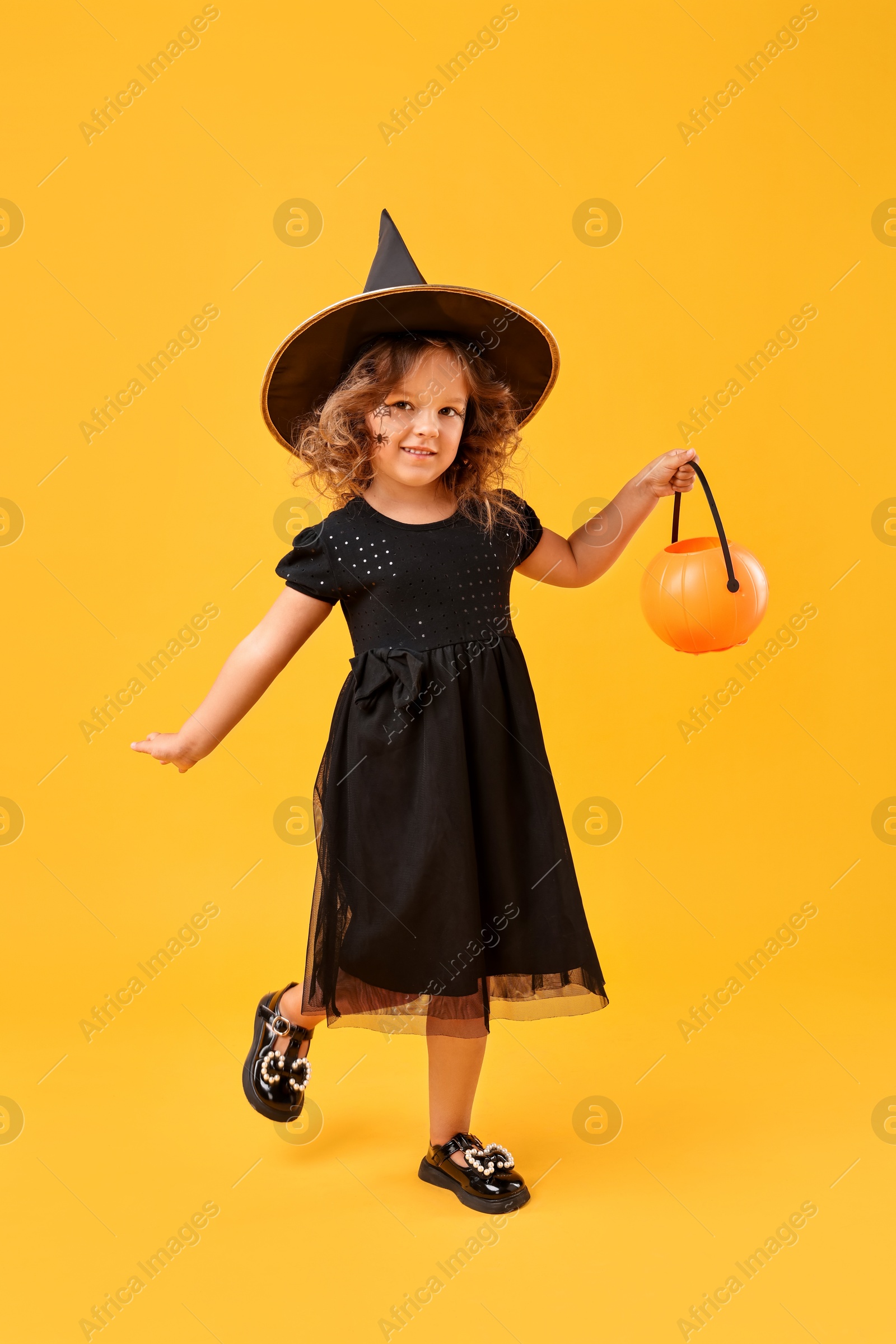 Photo of Cute girl with pumpkin bucket dressed like witch for Halloween celebration on yellow background