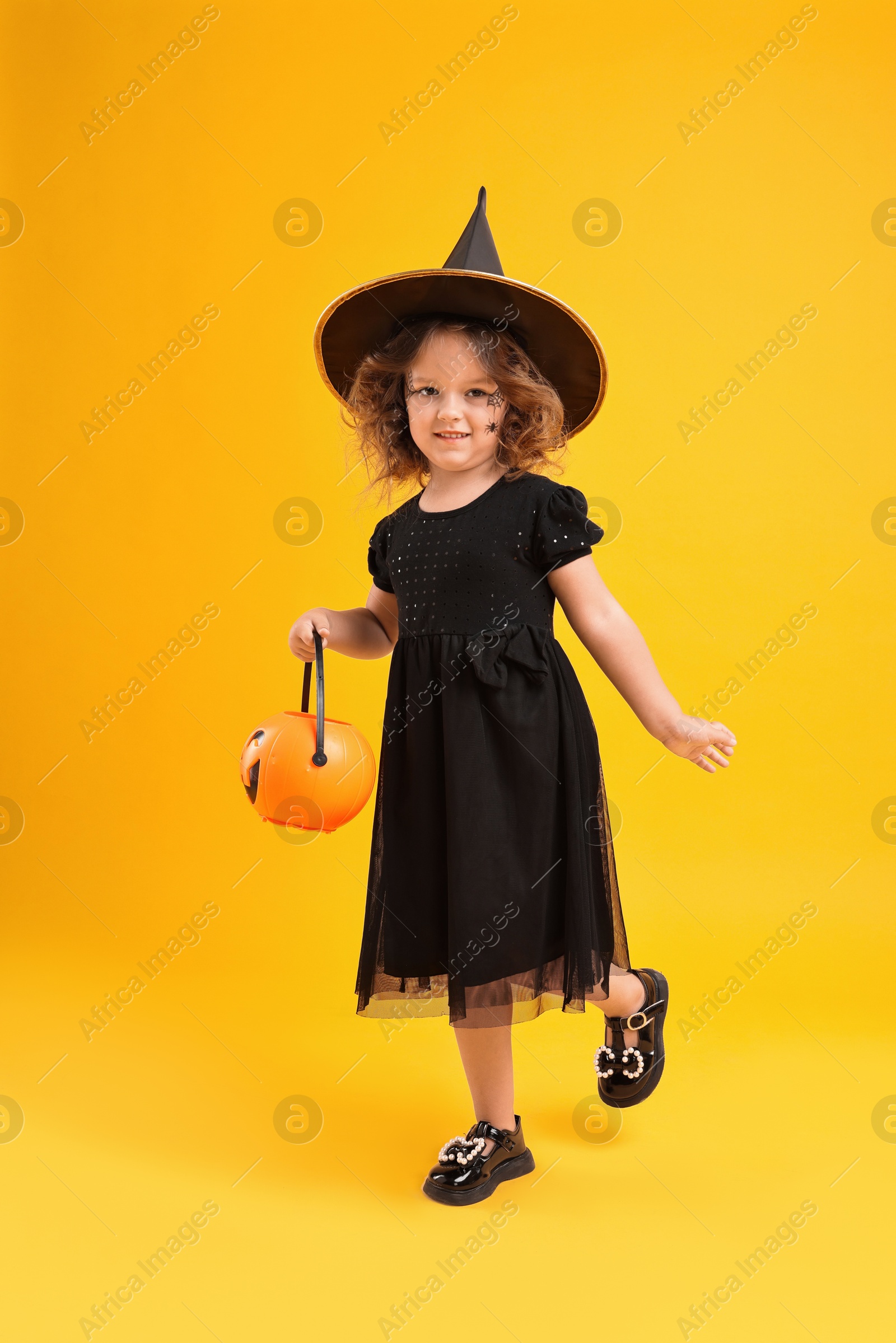 Photo of Cute girl with pumpkin bucket dressed like witch for Halloween celebration on yellow background