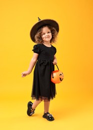 Cute girl with pumpkin bucket dressed like witch for Halloween celebration on yellow background