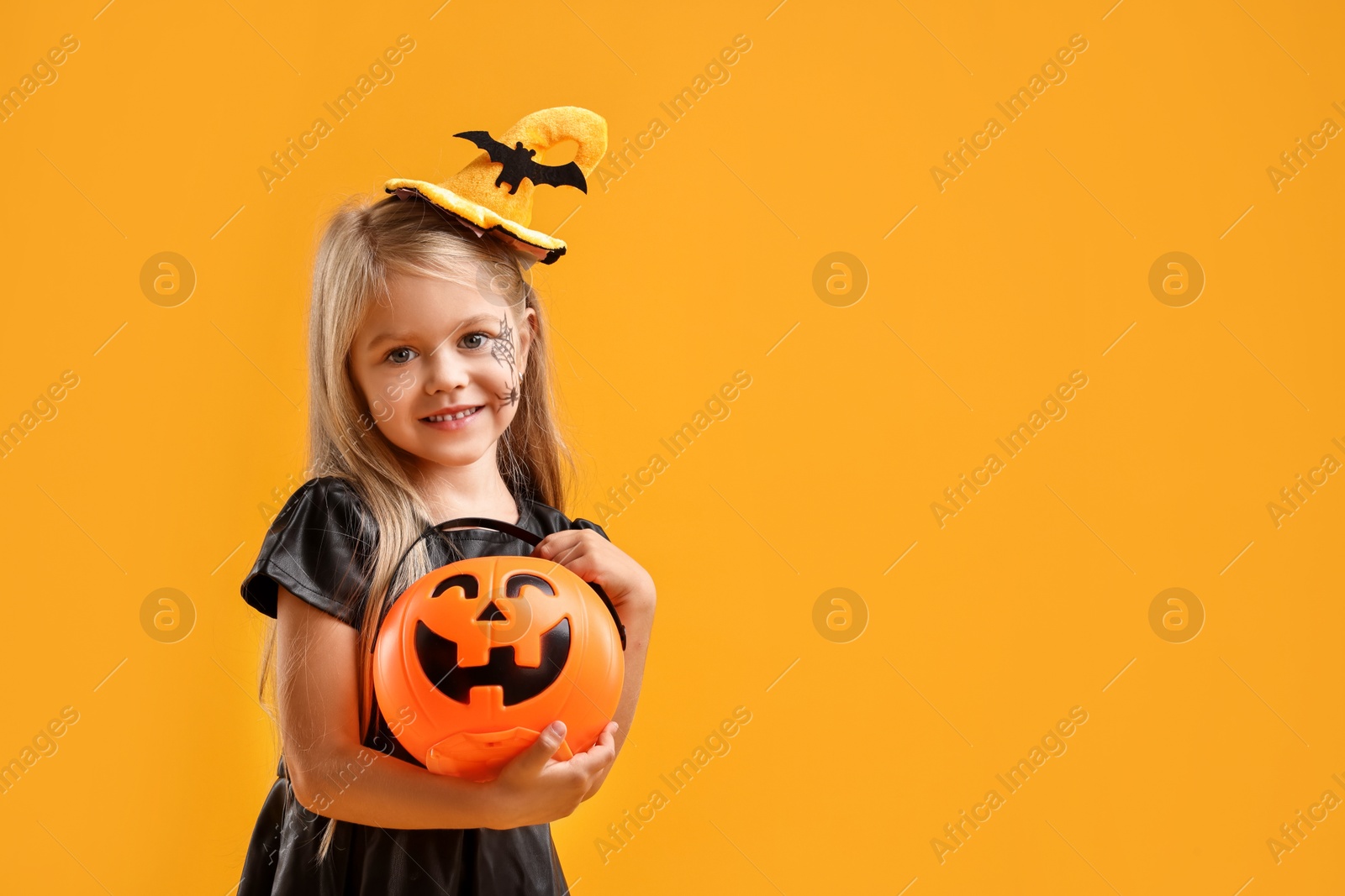 Photo of Cute girl with pumpkin bucket dressed like witch on yellow background, space for text. Halloween celebration