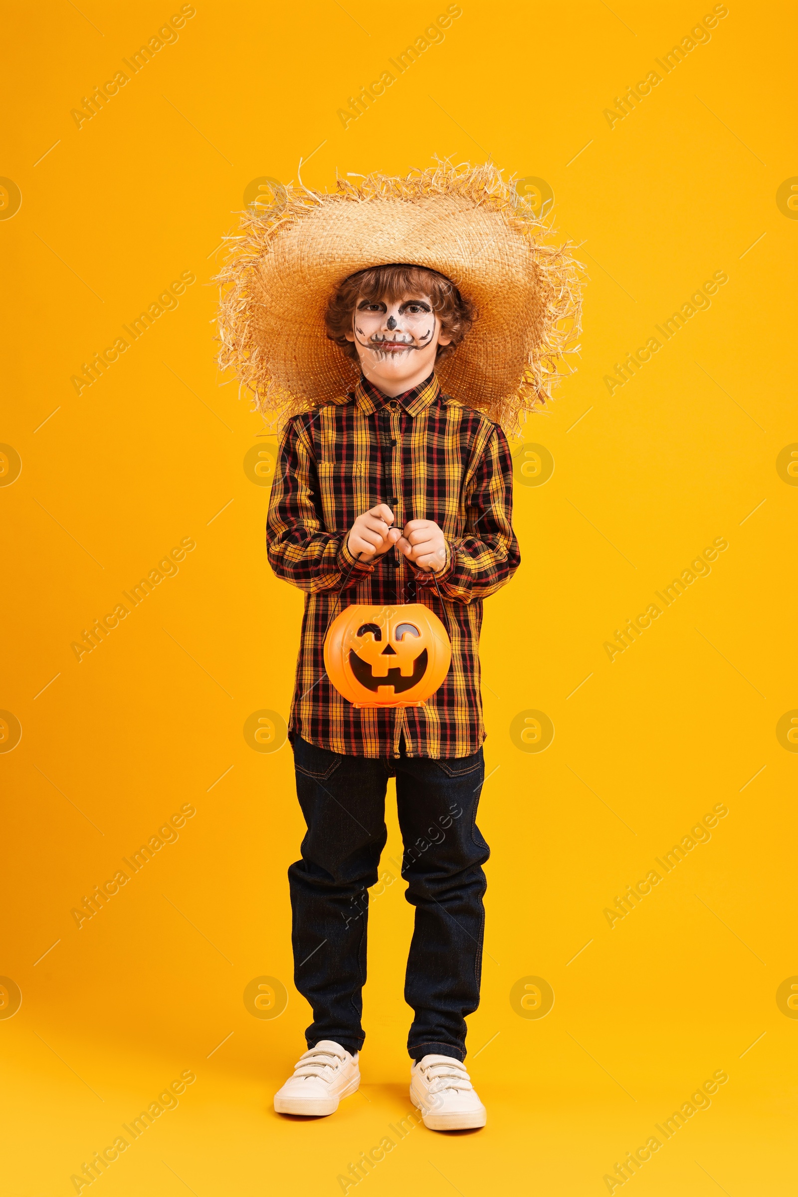 Photo of Funny boy with pumpkin bucket dressed like scarecrow on yellow background. Halloween celebration