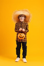 Photo of Funny boy with pumpkin bucket dressed like scarecrow on yellow background. Halloween celebration