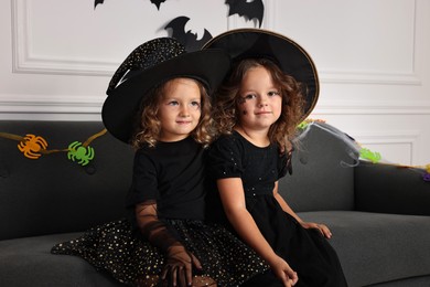 Photo of Cute girls wearing costumes on sofa indoors. Halloween celebration