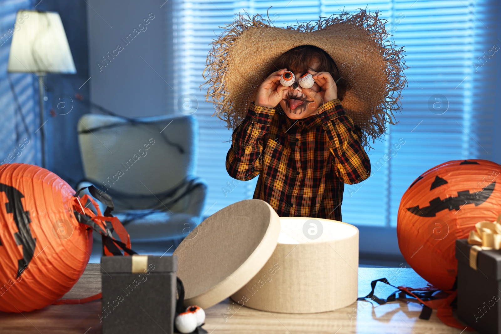 Photo of Cute boy dressed like scarecrow with decorative eyeballs, festive decor and gift boxes indoors at night. Halloween celebration