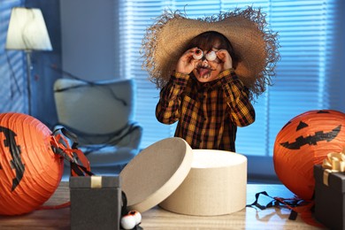 Cute boy dressed like scarecrow with decorative eyeballs, festive decor and gift boxes indoors at night. Halloween celebration