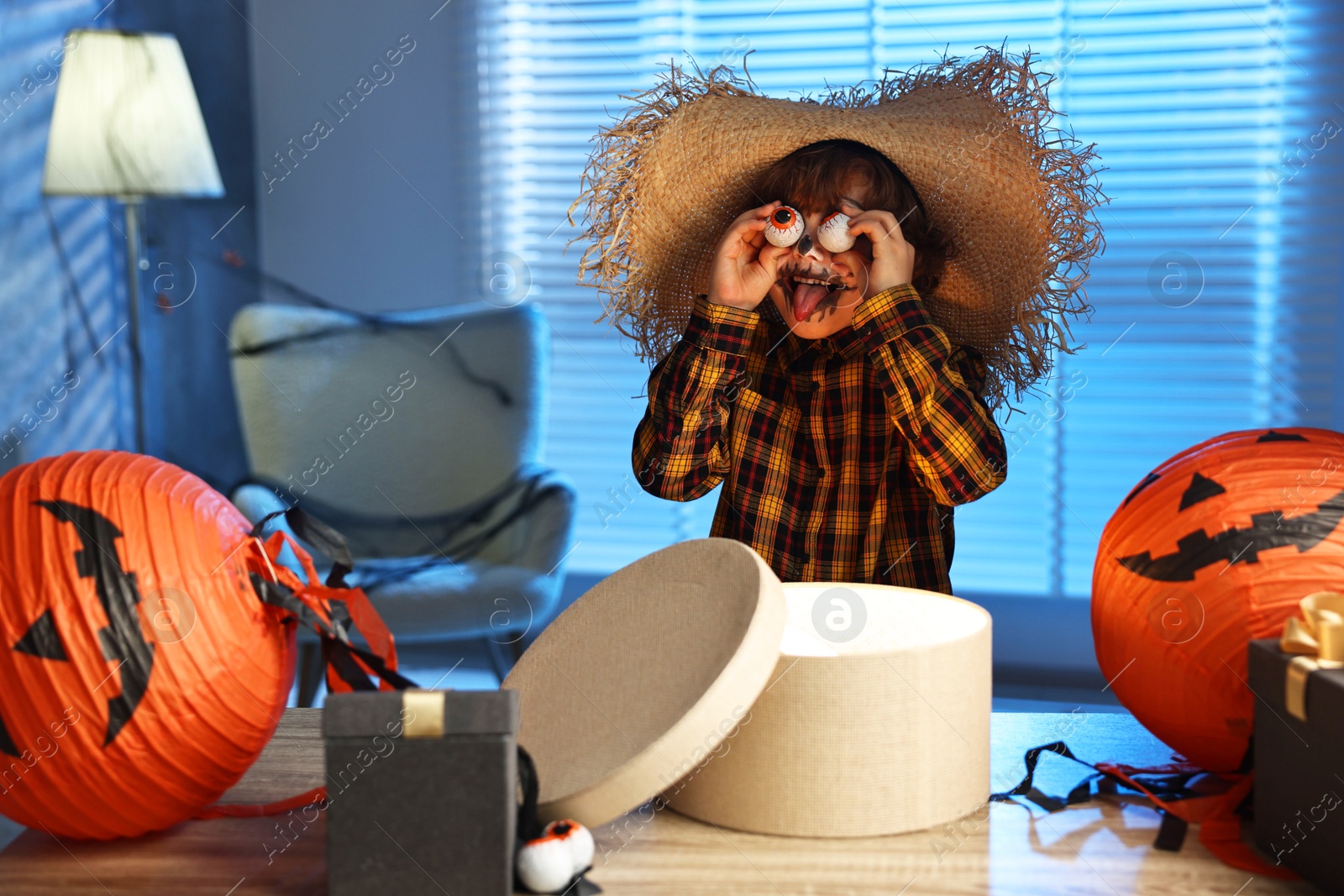 Photo of Cute boy dressed like scarecrow with decorative eyeballs, festive decor and gift boxes indoors at night. Halloween celebration