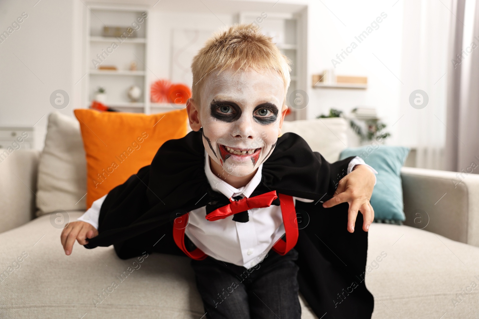 Photo of Funny boy dressed like vampire for Halloween celebration on sofa in room