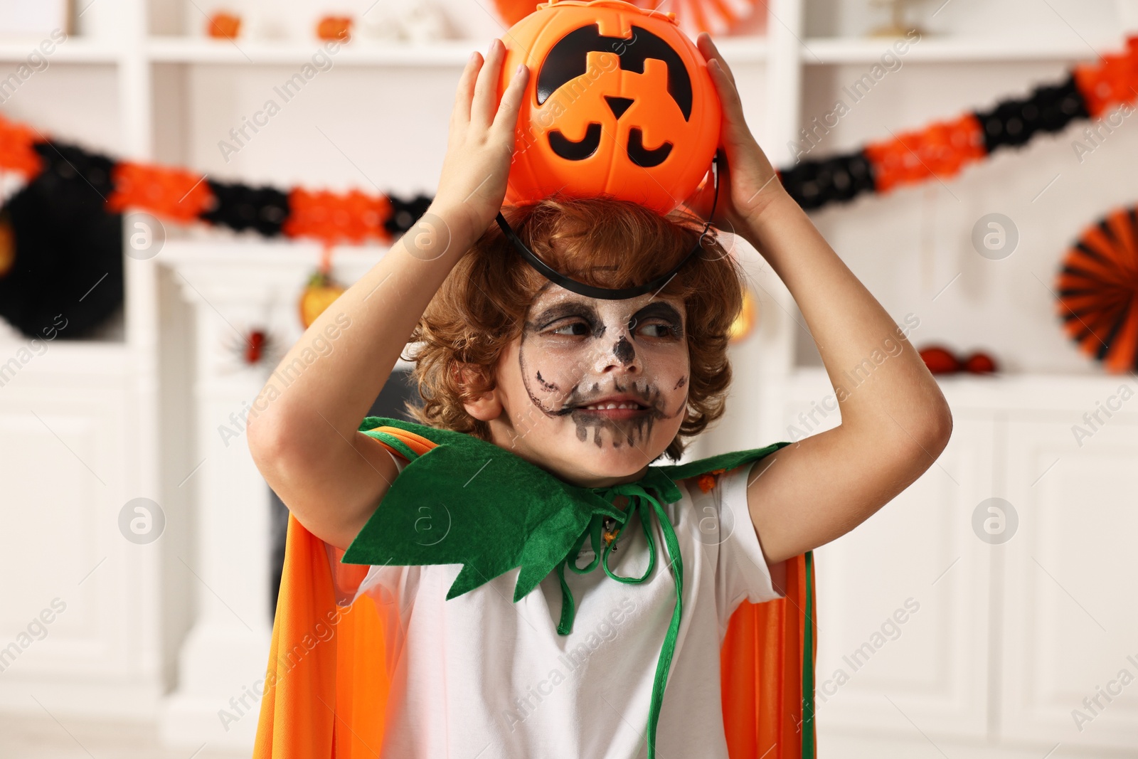 Photo of Funny boy with bucket dressed like pumpkin for Halloween celebration in room