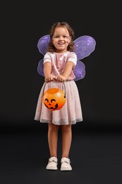 Photo of Cute girl with pumpkin bucket dressed like fairy for Halloween celebration on black background