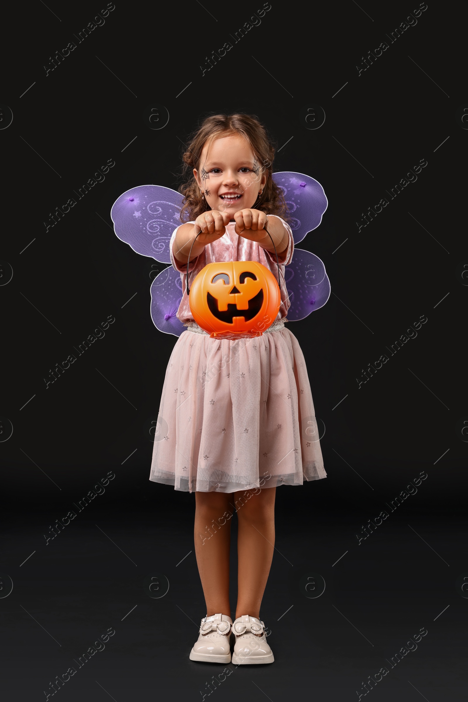 Photo of Cute girl with pumpkin bucket dressed like fairy for Halloween celebration on black background