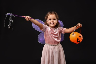 Photo of Cute girl with pumpkin bucket and magic wand dressed like fairy for Halloween celebration on black background