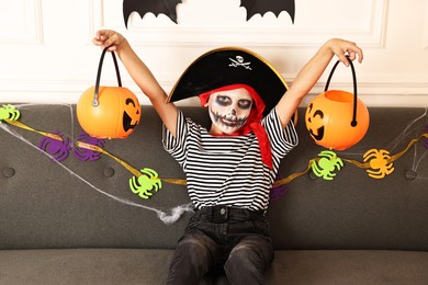 Funny boy with different Halloween decor dressed like pirate on sofa indoors