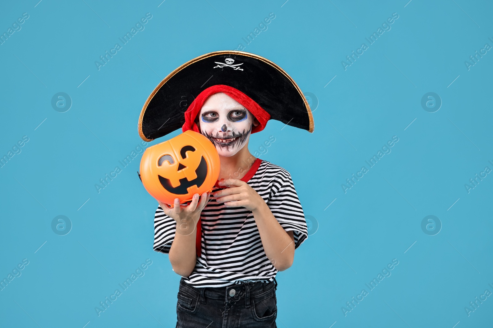 Photo of Funny boy with pumpkin bucket dressed like pirate on light blue background. Halloween costume