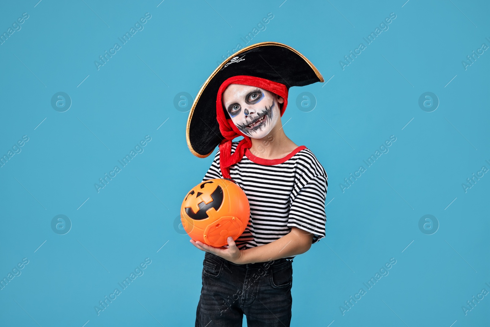 Photo of Funny boy with pumpkin bucket dressed like pirate on light blue background. Halloween costume