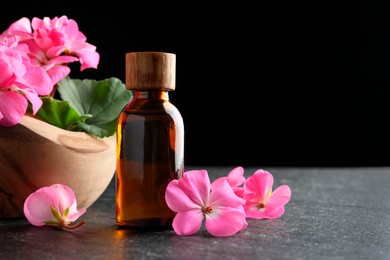 Photo of Bottle of geranium essential oil and beautiful flowers on black table, closeup. Space for text